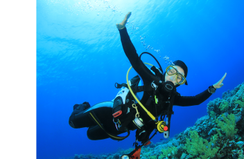 Promotional banner for Coral Adventures Netrani Murdeshwar featuring a scuba diver underwater with arms outstretched, wearing a black wetsuit, yellow diving mask, and oxygen tank. The background showcases a vibrant coral reef with clear blue water. On the left side, bold black text reads 'LEARN TO LIVE! LEARN TO DIVE!' against a white curved background with a coral illustration. The Coral Adventures logo is placed in the bottom right corner